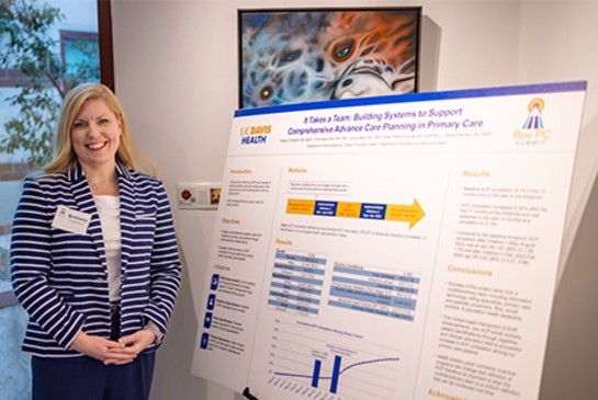 A woman with blond hair stands next to a research poster that highlights teamwork to support advance care planning in primary care.